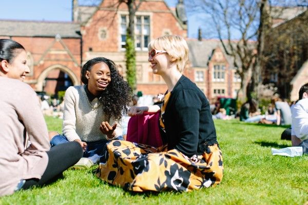Three students are smiling and talking, sitting on the grass in front of the Clothworkers' Building on a sunny day.