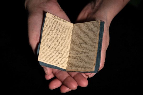 Open hands holding a tiny book filled with handwriting, against a black background.