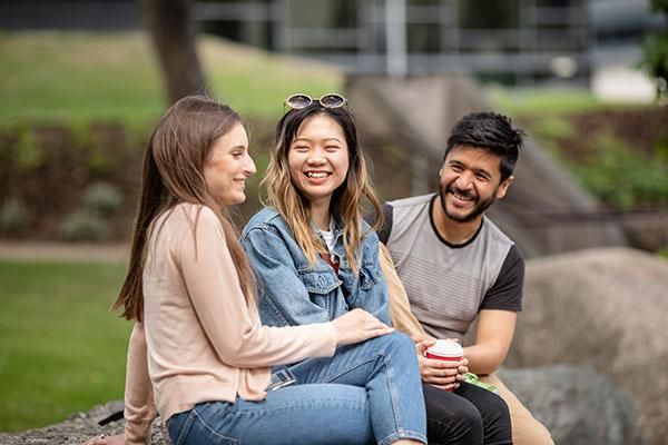 Three international students laughing whilst sitting on a rock on campus
