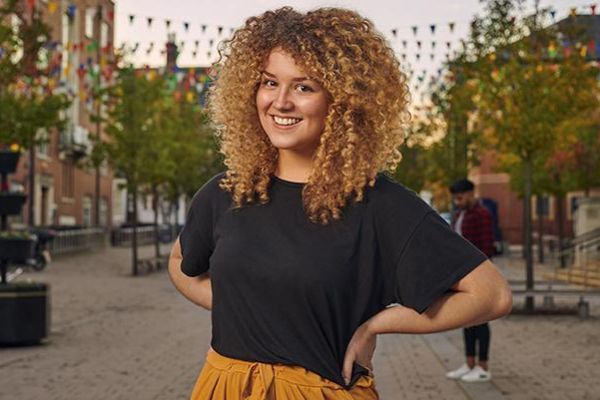 A student with curly hair, a black t-shirt and orange trousers is standing outside Leeds University Union. There are trees and colourful flags in the background.