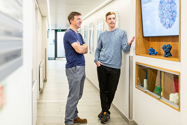 A male student and his research supervisor standing in front of a monitor attached to a wall discussing work