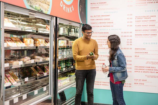 Students chatting while choosing drinks from a large cafe fridge stocked with sandwiches and drinks