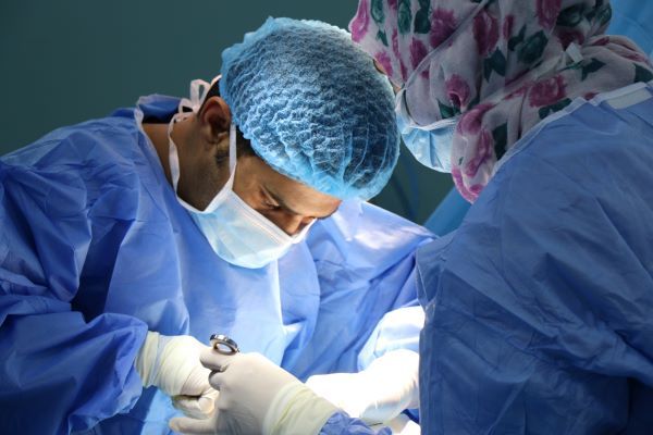 A surgeon in blue scrubs, blue cap and white mask performs surgery under a bright life. His assistant, with her back to the camera, is wearing blue scrubs, white mask and a pink floral pattern headscarf,