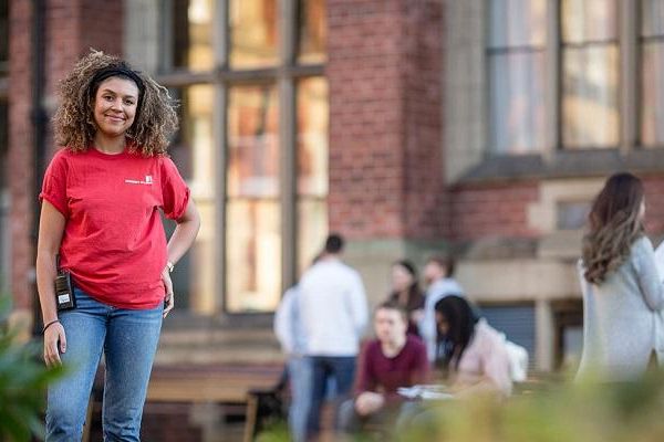 A student ambassador stood in front of an old building on campus during open day, smiling at the camera. People are looking round in the background.