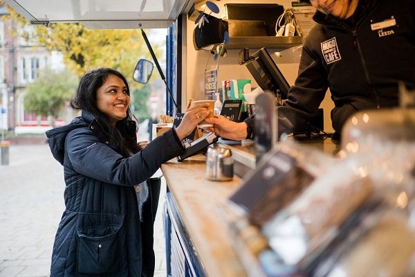 A student buying a coffee at a coffee stall on a cool, fresh day on campus and smiling at the vendor.