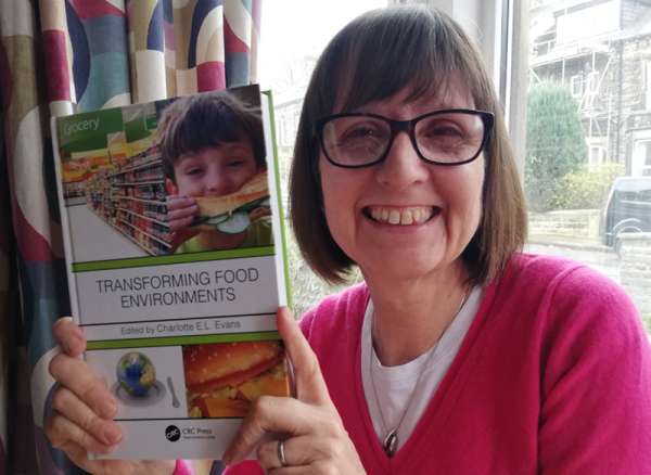 A person smiling and holding a copy of Transforming Food Environments, a book edited by Dr Charlotte Evans.