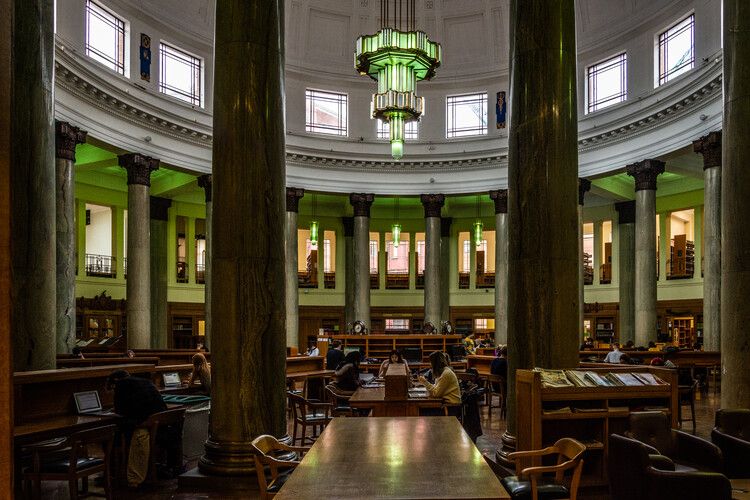 Students study at wooden tables in the Brotherton Library. The large domed ceiling with an art deco light fitting can be seen as well as marble columns.