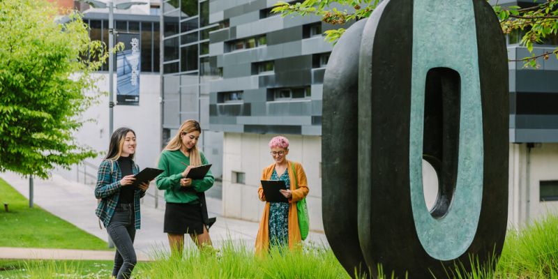 Three students with clipboards study a large bronze Barbara Hepworth bronze artwork which stands outside on campus.