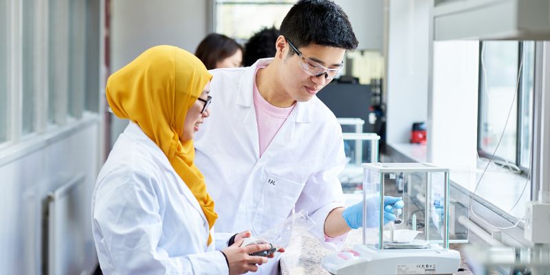 Two students in lab coats work with specialised equipment.