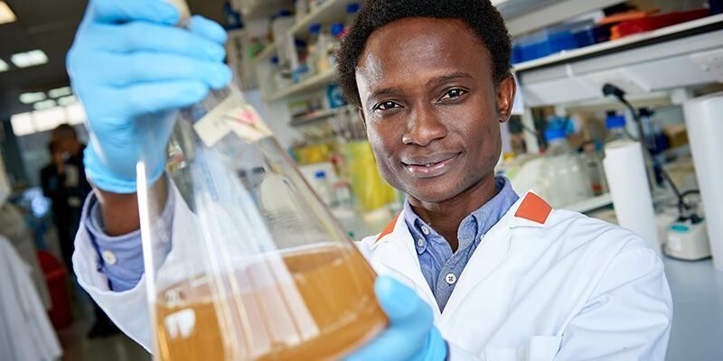 Dr Adeyemi wearing a white lab coat and blue gloves holding a beaker filled with liquid.