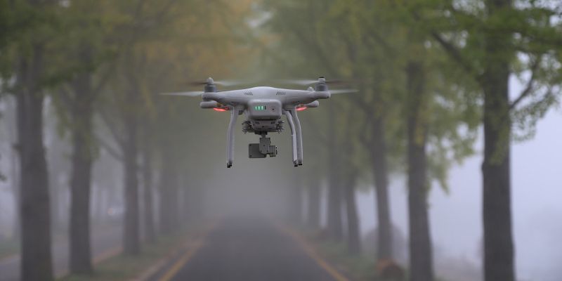 A silver drone flies above a road lined by trees on a foggy day