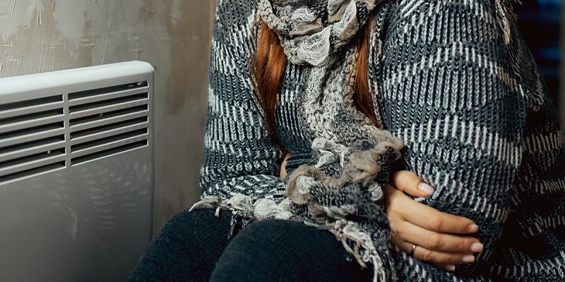 Person in thick jumper and scarf sits close to a small radiator hugging themselves to keep warm.