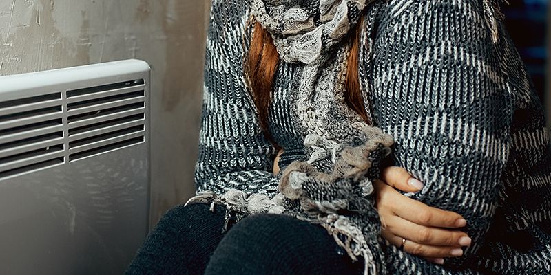 Person in thick jumper and scarf sits close to small radiator while hugging themselves to try to keep warm.