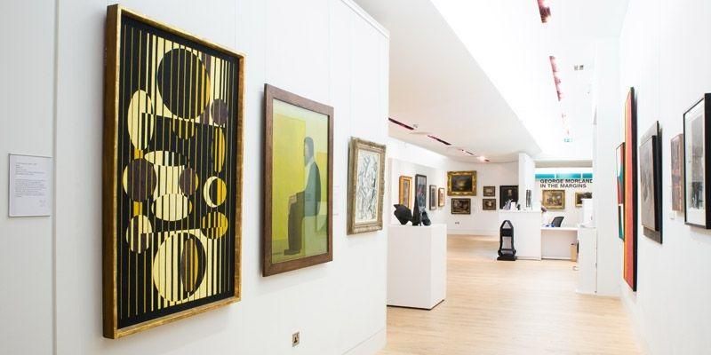 An art gallery with walls of paintings on either side. One painting is black with yellow circles and another is yellow with an image of a person sat on a chair.