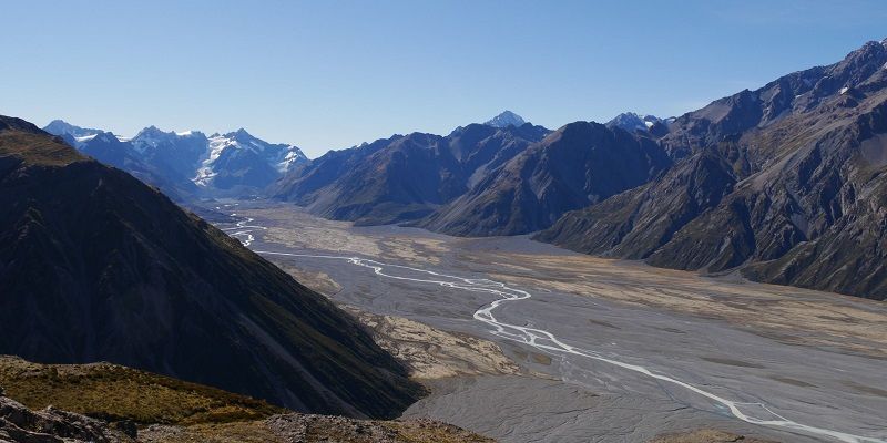 A wide flat-bottomed valley with a narrow winding river in it, dwarfed by steep rocky mountains on either side. In the background, there is a glacier