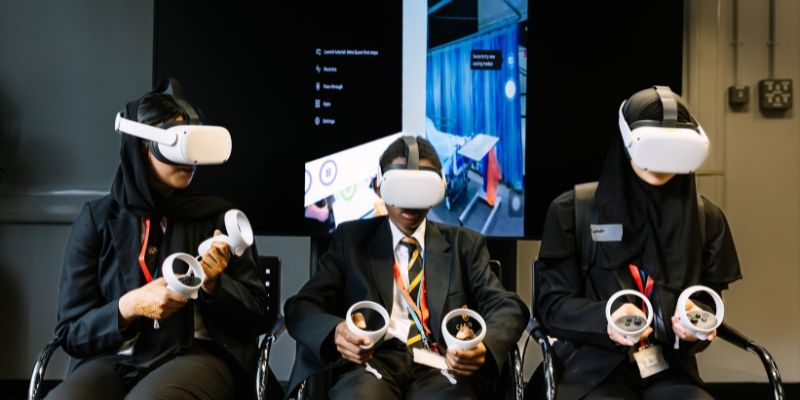 Three high school pupils wearing VR headsets and using VR hand controls, seated in front of a screen showing a hospital scene