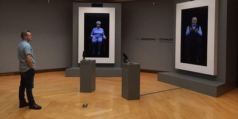 A person standing in a gallery looking at two large screens, each screen showing an image of a person on them. There are two black plinths between the screens and the person.