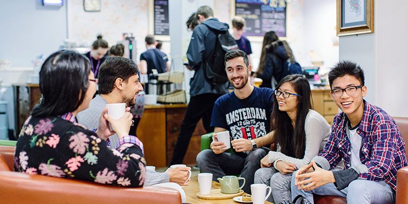 A group of international students sat around a coffee table in a cafe, chatting and enjoying drinks