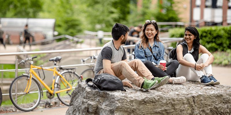 Three students sat on a rock on campus. There is a yellow bike in the background.