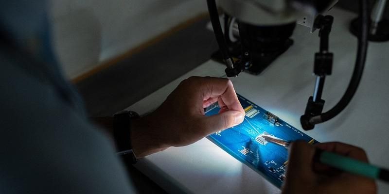 The image shows an engineering soldering wire onto a circuit board. He is using a magnifying system to guid his movements.