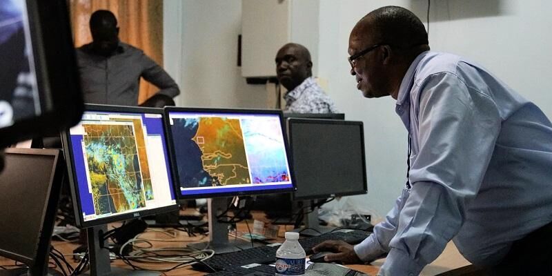 The image shows a eather forcaster looking at satellite images on a screen. There are other forecasters in the office.