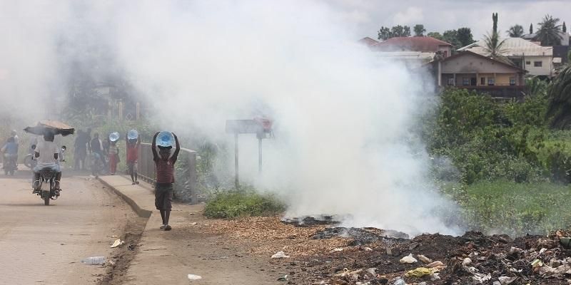 Picture shows a smouldering fire by the side of a road in Africa. People are having to walk through a cloud of blue smoke