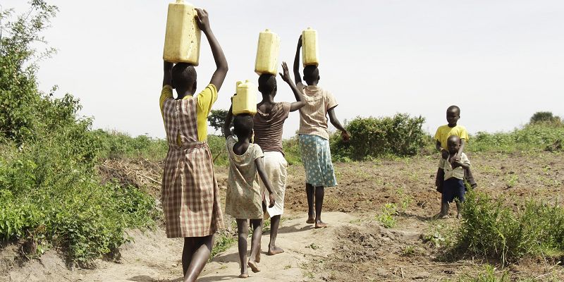 The image shows a group of people carrying containers of water across a field.