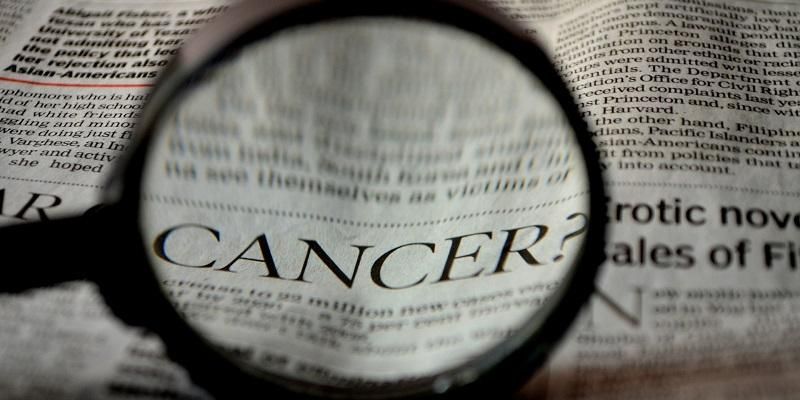 The image shows a magnifying glass over a newspaper. The word that is magnified is - Cancer,