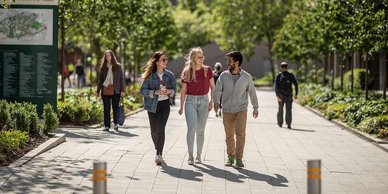 Three students exploring the University of Leeds campus on a sunny day &mdash; surrounded by trees.