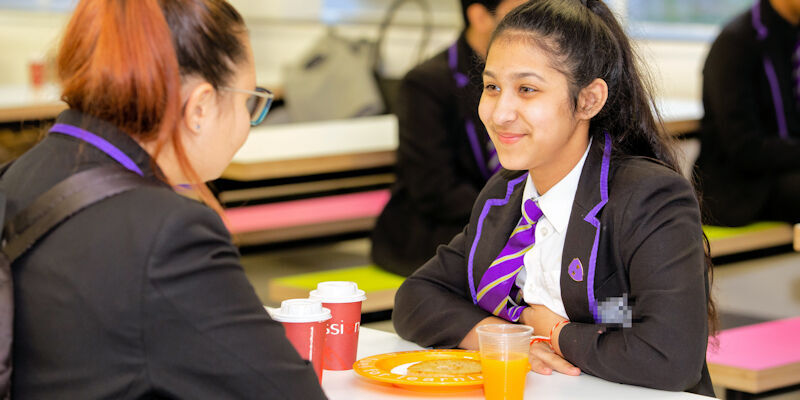 Two teenage girls sit eating breakfast at school in Leeds, wearing smart black blazers with purple ties.