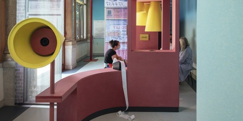People sat on pink benches with yellow cone-like sculptures in an indoor space with windows in the background