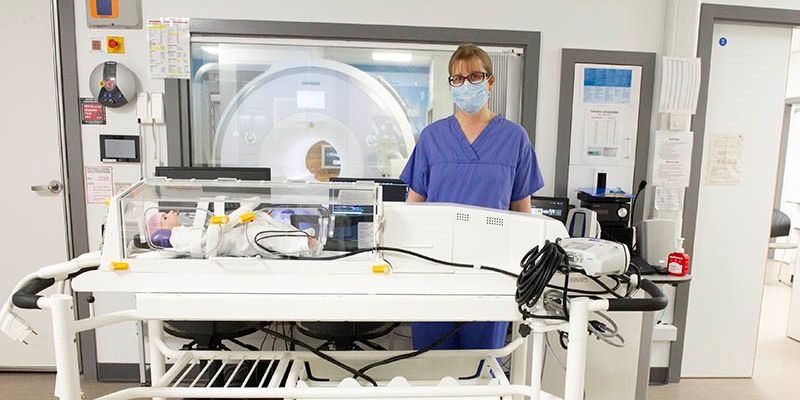 A medical professional in scrubs and face mask stood next to a piece of equipment designed to allow a baby to pass through a MRI scanner