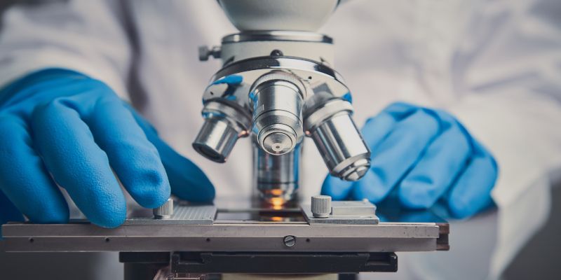 A close-up of a microscope and the hands of a researcher wearing blue gloves and a white coat.