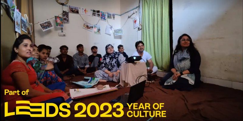 A community group in India viewing an artwork made by artificial intelligence. 11 people sit on the floor around a projector, looking at the artwork. Logo on the image says 'Part of Leeds 2023 year of culture'.