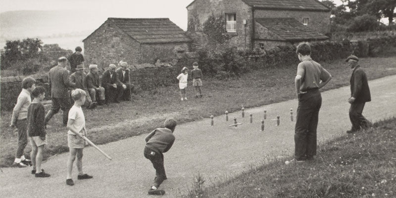 Children playing wallops (nine-pins) in the street at Castle Bolton in Wensleydale, North Yorkshire, 1964. A group of men can be seen sitting on a bench, watching men from the village playing quoits on the grass verge.