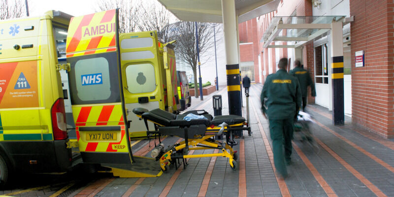 An ambulance parked outside Accident and Emergency (A&E) with its back doors open and a stretcher on wheels nearby, as two paramedics walk towards the A&E entrance.