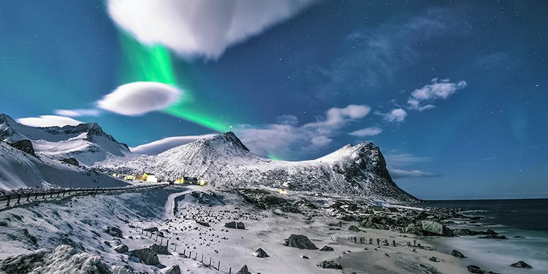Green streaks of aurora borealis among white clouds over snowy mountain peaks in the arctic.