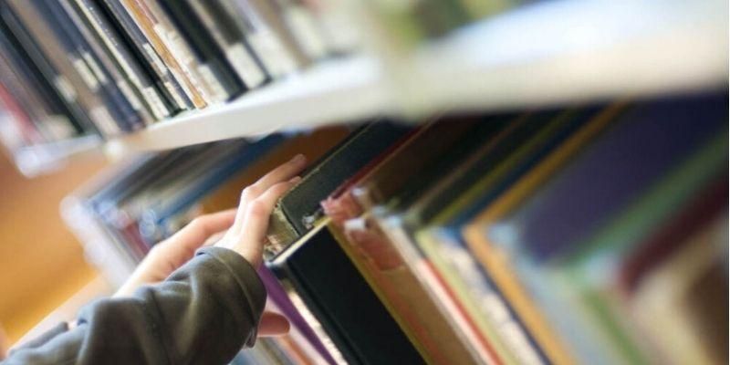 A pair of hands reaches for a book on a library bookshelf.