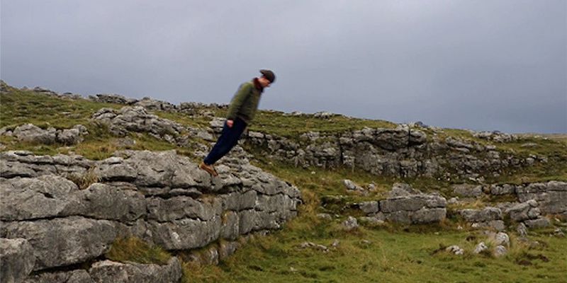 A person on the edge of a boulder on a hillside leaning forward as if about to fall off