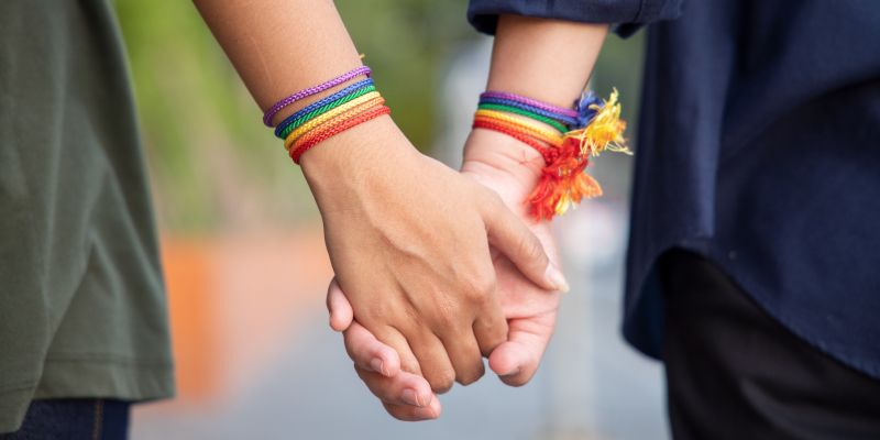 The lower arms and hands of an adult couple holding hands, both with rainbow coloured friendship bracelets.