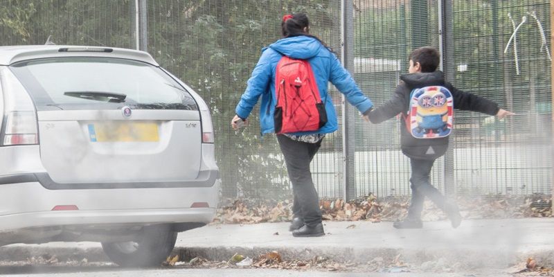 An adult and child holding hands, walking on a pavement with visible car fumes on the road next to them