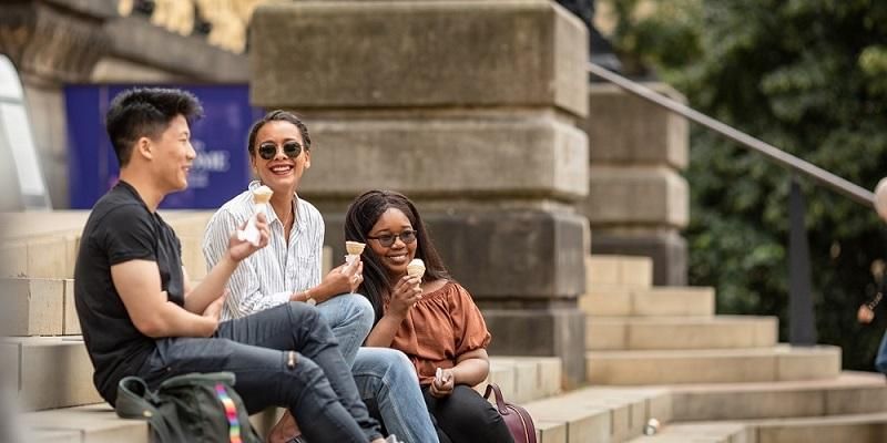 A small group of students sit on Leeds museum steps in the sunshine eating ice cream.