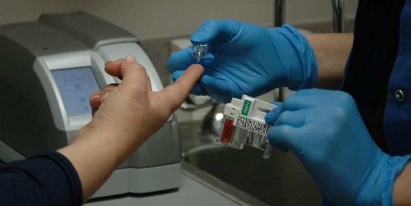 The image shows a patient having a finger-prick blood test for diabetes.